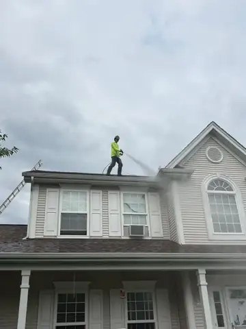 Worker in a yellow shirt spraying a grey shingle roof from the ridge
